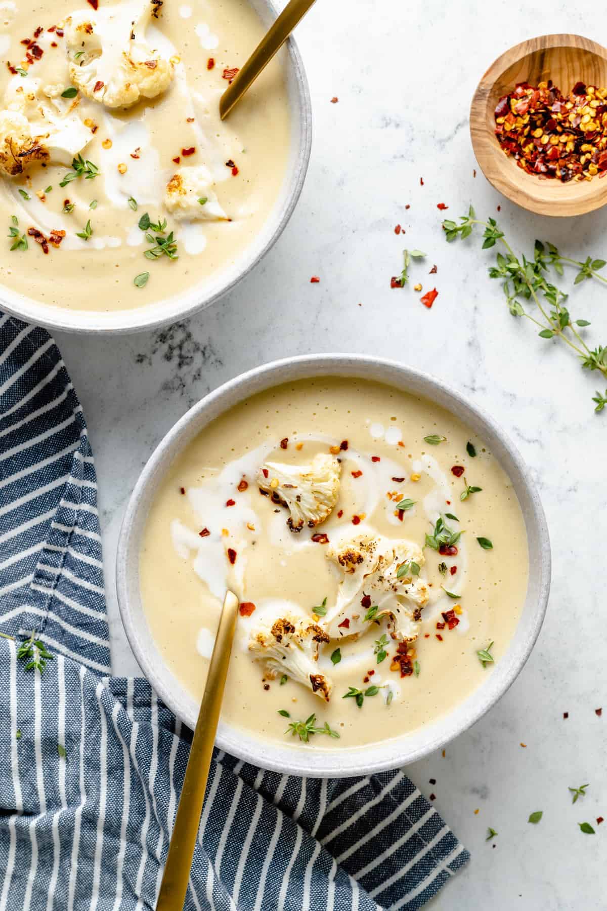 Overhead view of two bowls filled with cauliflower soup, garnished with red pepper flakes, thyme, and coconut milk