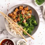 General Tso Tofu in bowl with rice and broccoli with chopsticks and garnishes