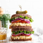 Two stacked vegan tuna sandwiches topped with sprouts, lettuce, red onion, and tomatoes
