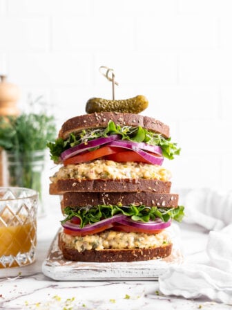 Two stacked vegan tuna sandwiches topped with sprouts, lettuce, red onion, and tomatoes