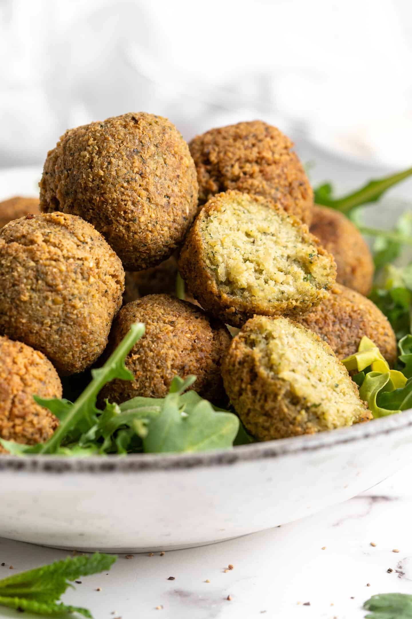 Closeup of falafel in bowl with greens, with one cut open to show fluffy inside