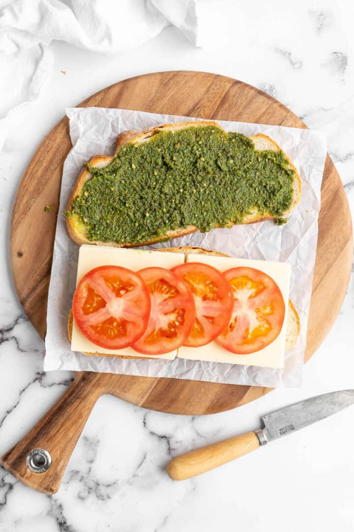 This vegan Caprese sandwich layers plant-based mozzarella with fresh tomatoes and parsley pesto, all on crusty French bread. Best lunch ever! One slice of bread spread with pesto, another topped with mozzarella and tomatoes on round cutting board