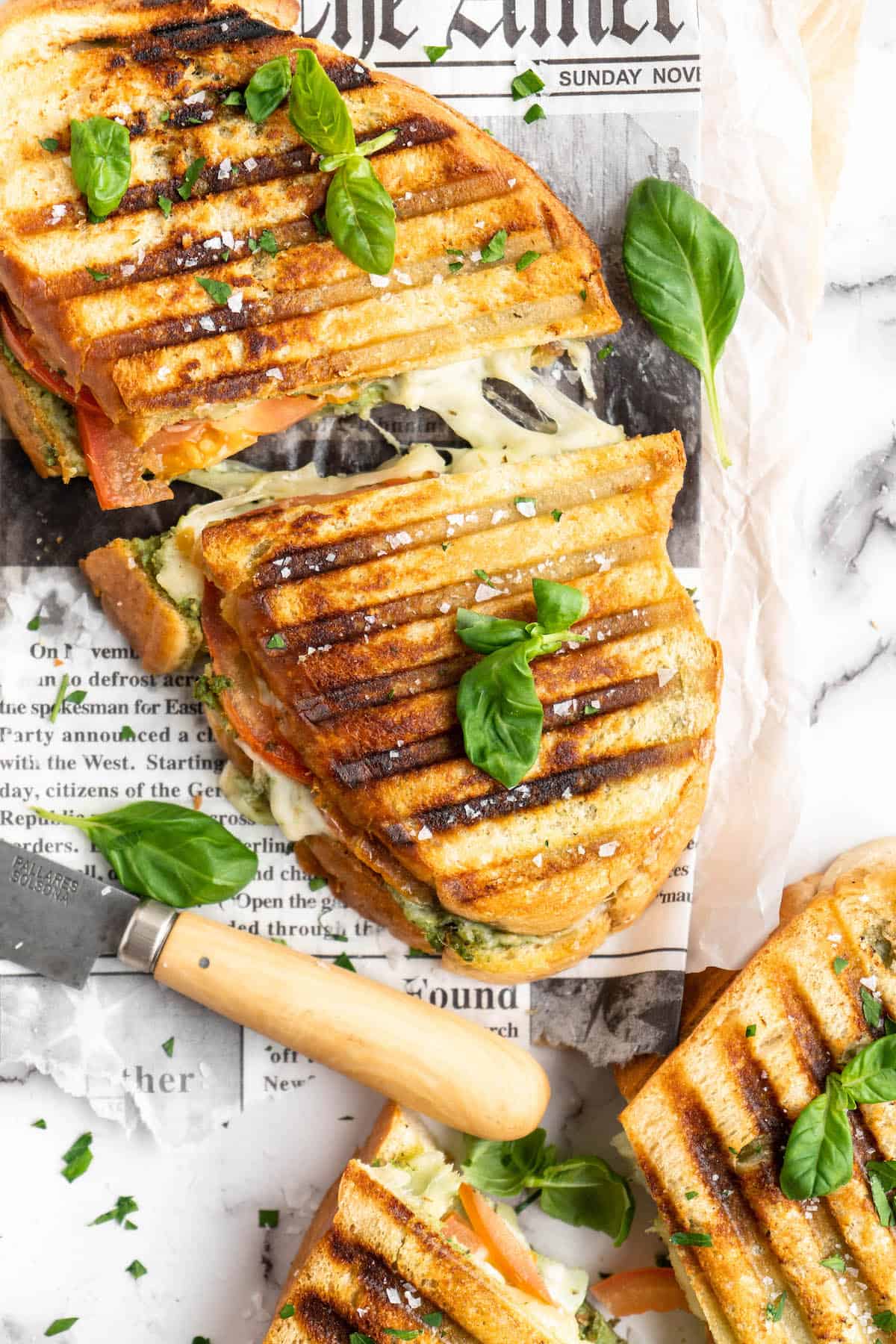 Halved vegan caprese sandwiches set on newspaper, with basil leaves scattered over them for garnish