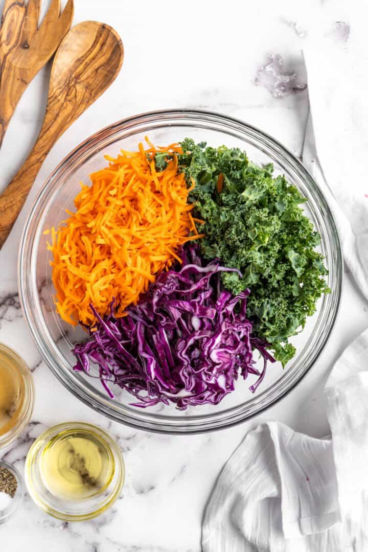 Overhead view of kale slaw ingredients added to a large glass mixing bowl.