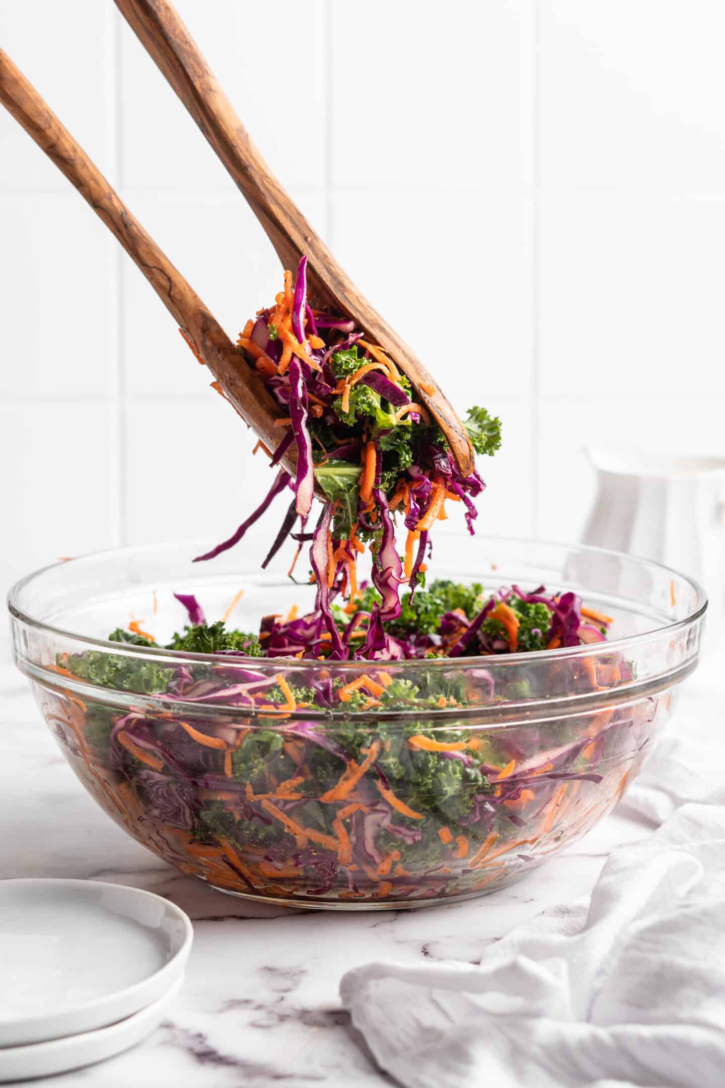 Salad tongs lifting kale slaw from a glass mixing bowl.