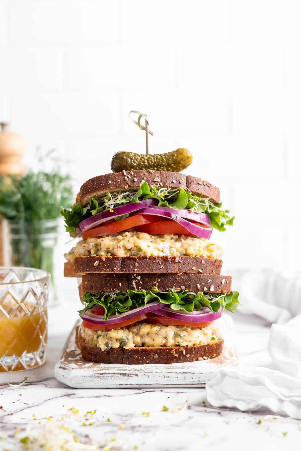 Two stacked vegan tuna sandwiches topped with sprouts, lettuce, red onion, and tomatoes