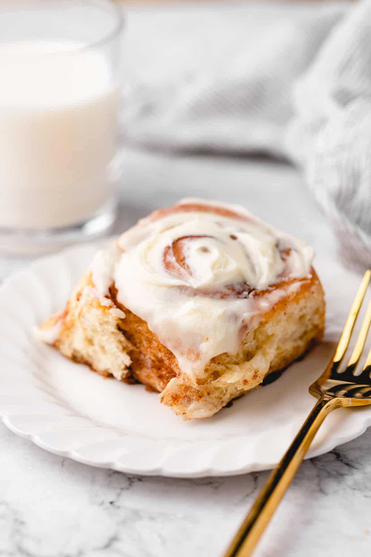 Vegan cinnamon roll on white plate with gold fork, with glass of milk in background