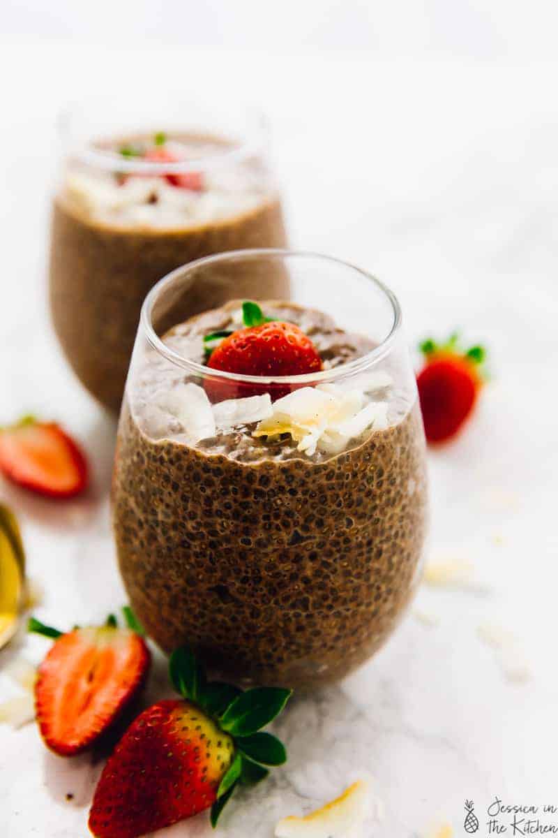 A wine glass filled with chocolate chia pudding, with coconut flakes and a strawberry on top, and cut up strawberries in the background