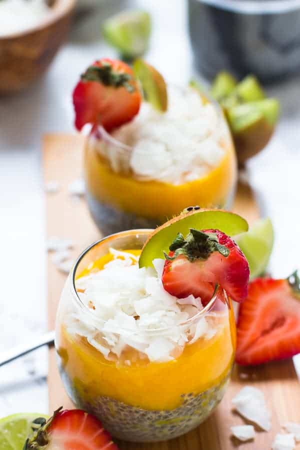 A wine glass with chia seeds in the bottom, mango pudding on top, and topped with coconut flakes, with a strawberry and kiwi slice on the edge of the glass, a glass of pudding in the background, and strawberries and kiwis on the table