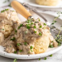 A plate with two biscuits and gravy on it, garnished with chives, with a fork and knife on the plate and biscuits in the background