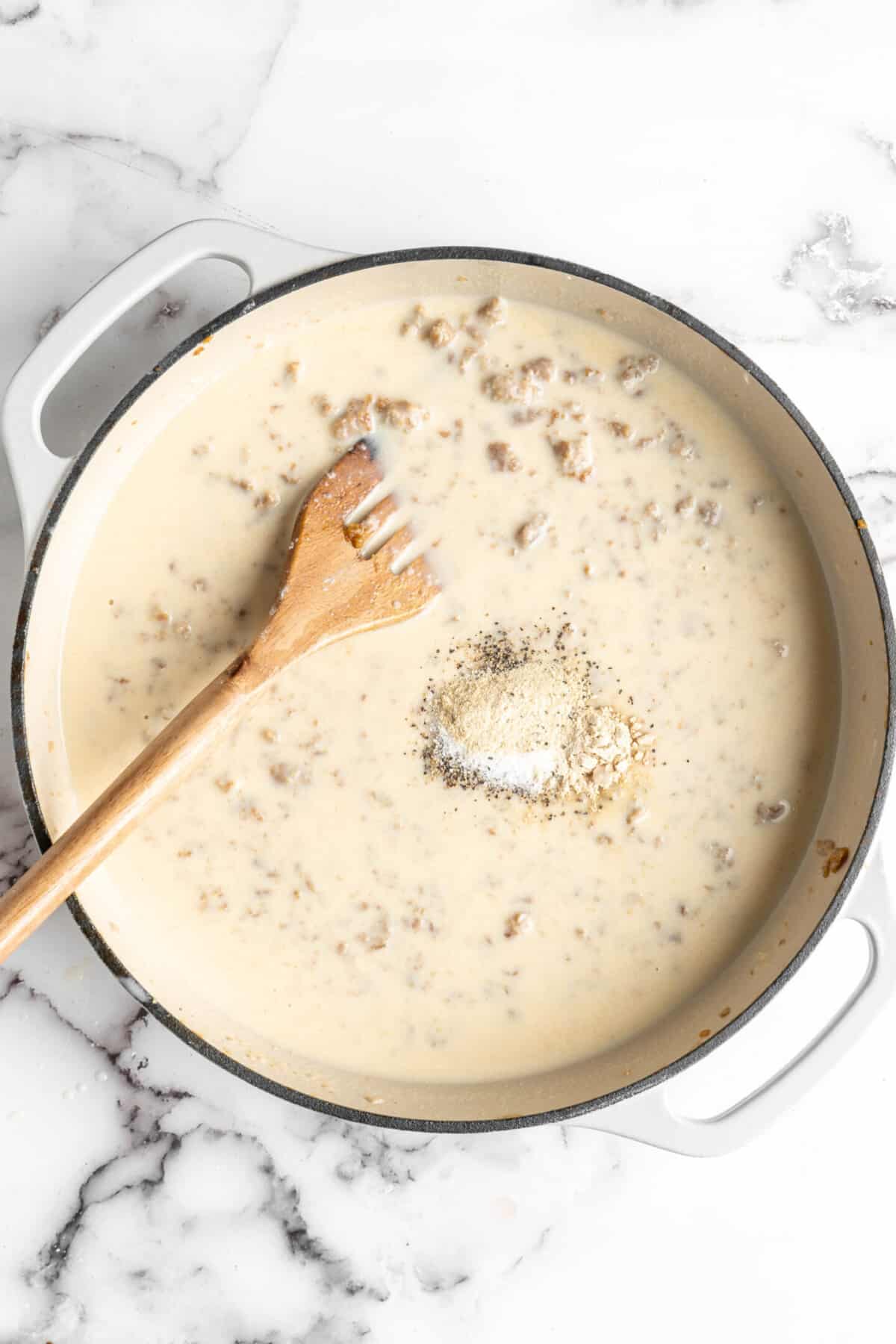 A very wet gravy in a dutch oven with a pile of seasonings on top, and a slotted wooden spoon