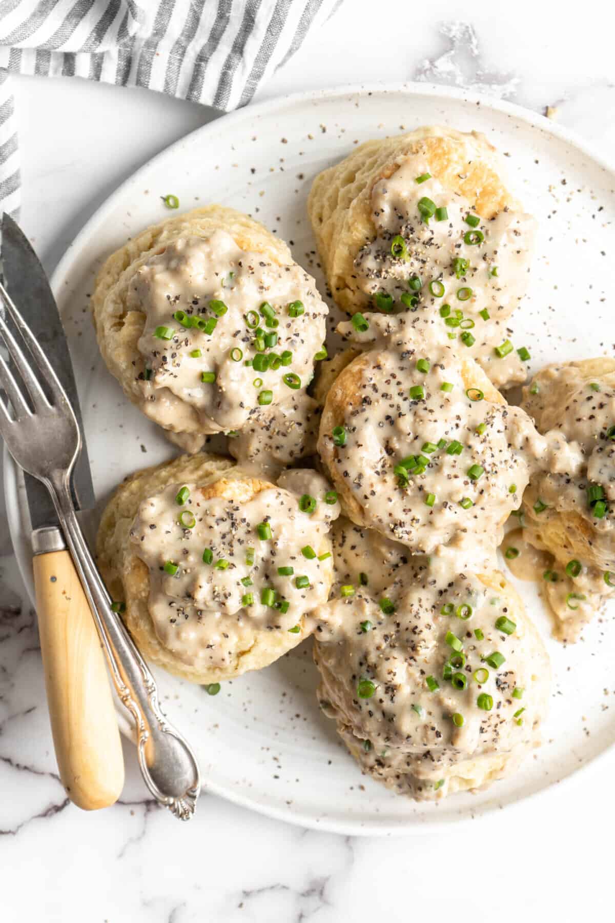 A plate with six biscuits covered in gravy and chives, with a fork and a knife