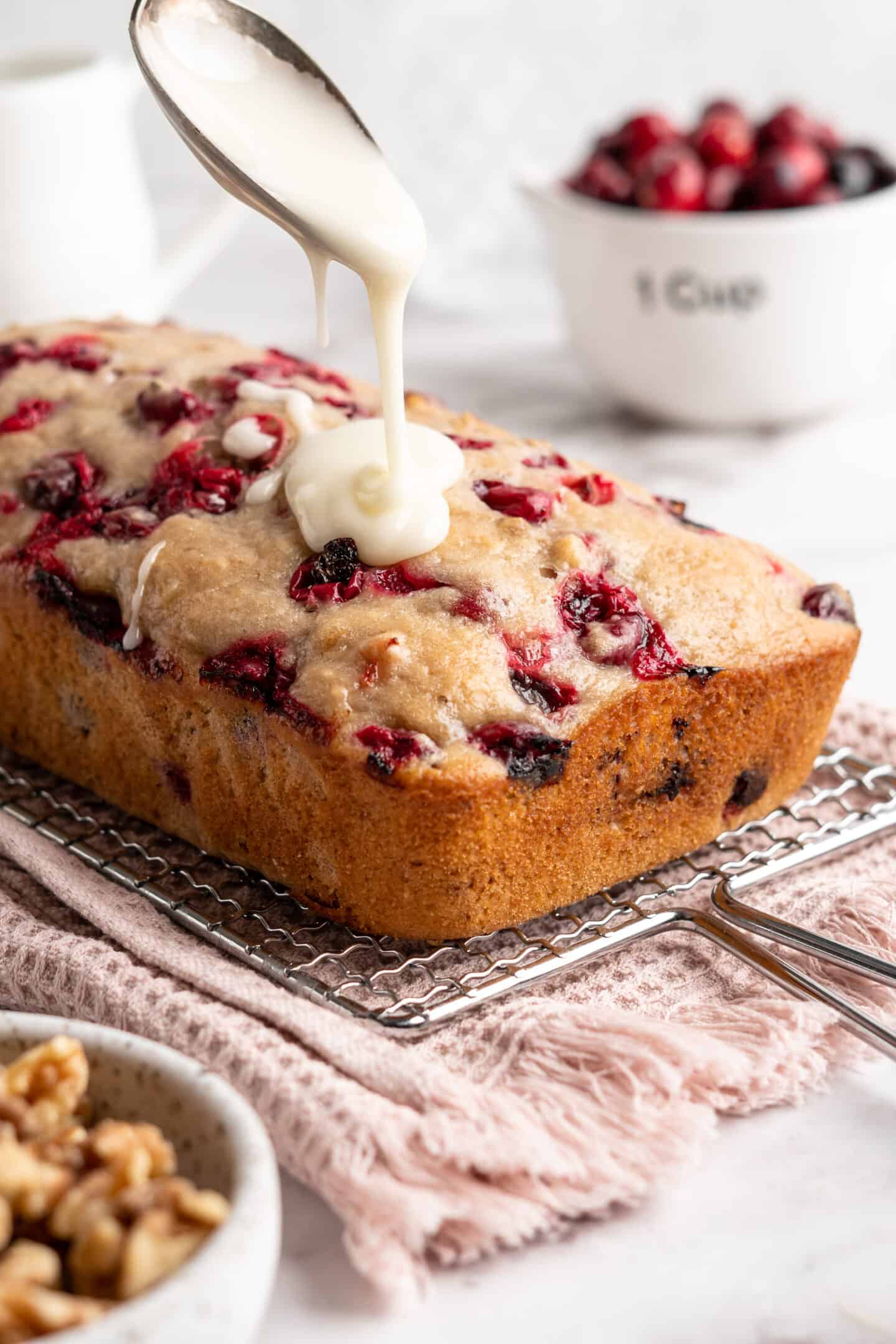 A loaf of cranberry walnut bread on a cooling rack, with glaze being drizzled on top.