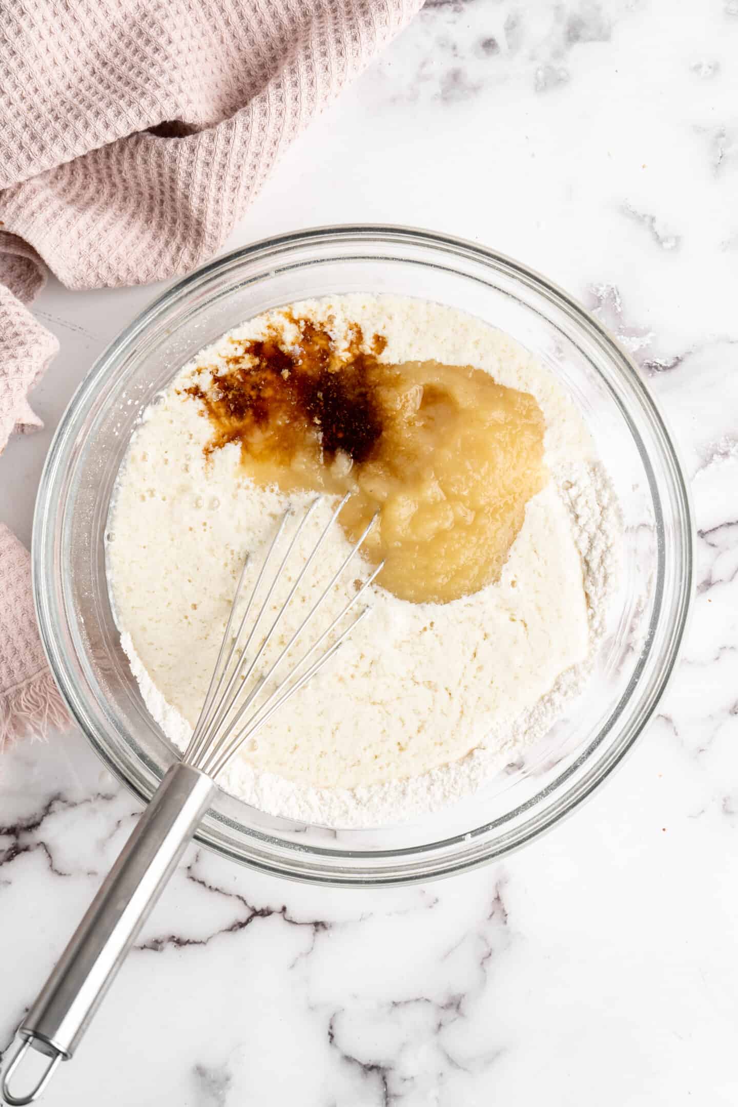 A mixing bowl with a flour and sugar mixture, topped with applesauce, vegan buttermilk, and vanilla extract, with a whisk in the bowl