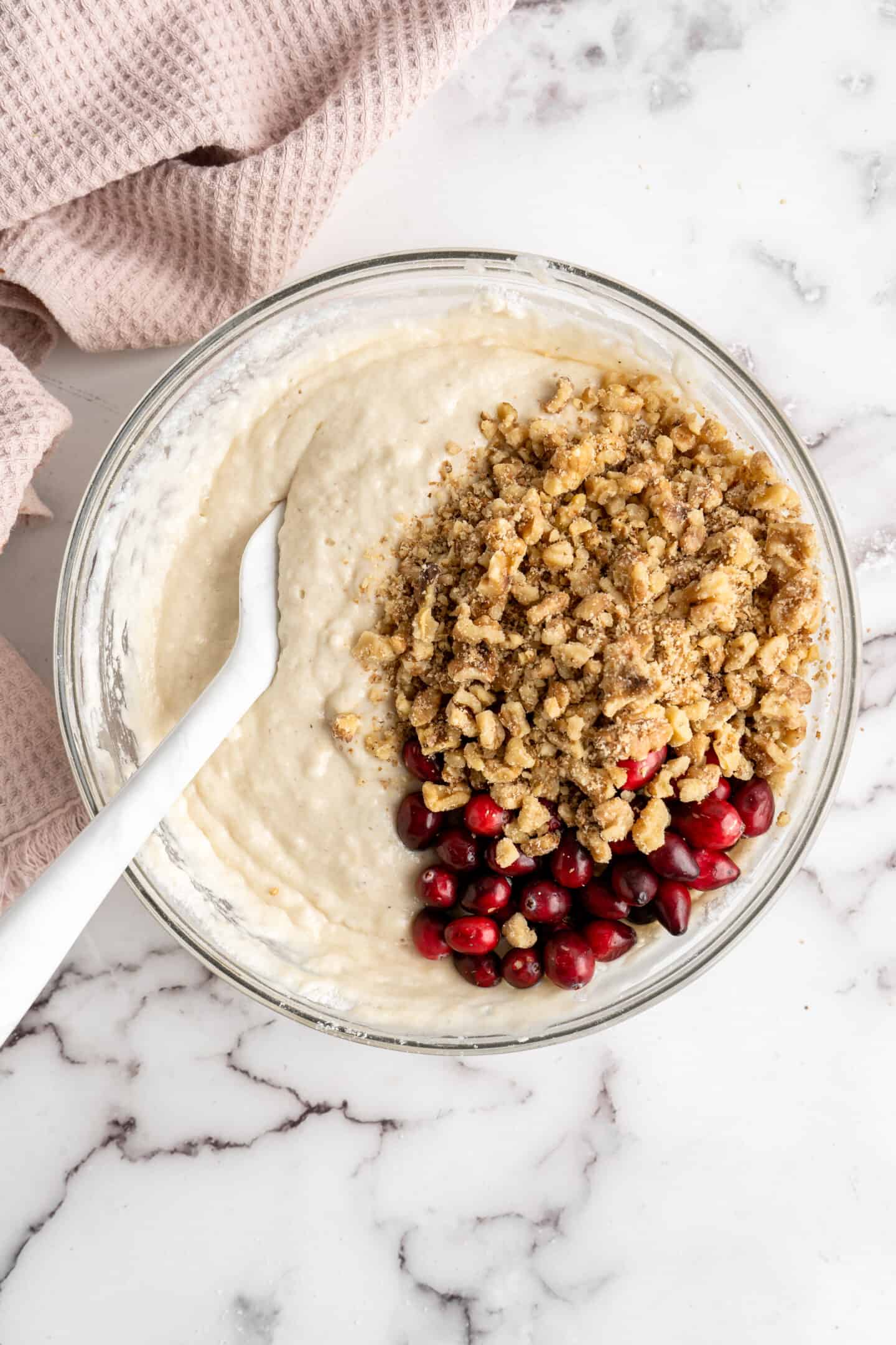 A mixing bowl with walnuts and cranberries sitting on top of cranberry walnut bread batter, with a spatula in the bowl