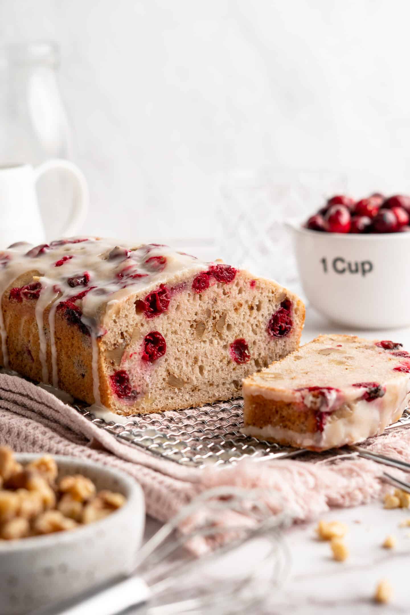 A loaf of cranberry walnut bread on a cooling rack, sliced into, with a slice of bread in front of it and a bowl of cranberries behind it