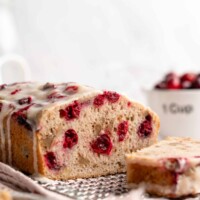 A loaf of cranberry walnut bread on a cooling rack, sliced into, with a slice of bread in front of it and a bowl of cranberries behind it