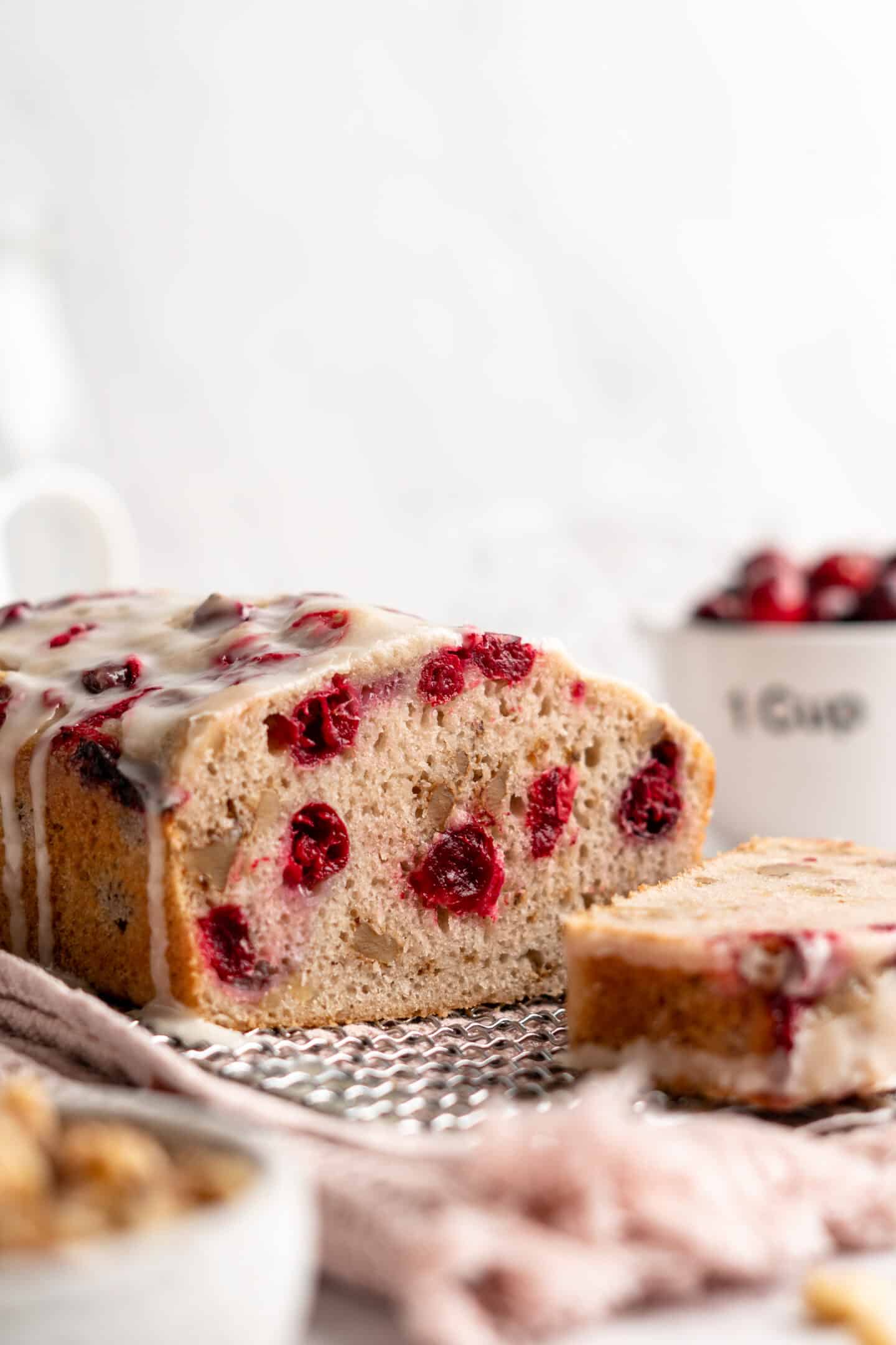 A loaf of cranberry orange bread on a cooling rack, sliced into, with a slice of bread in front of it and a bowl of cranberries behind it