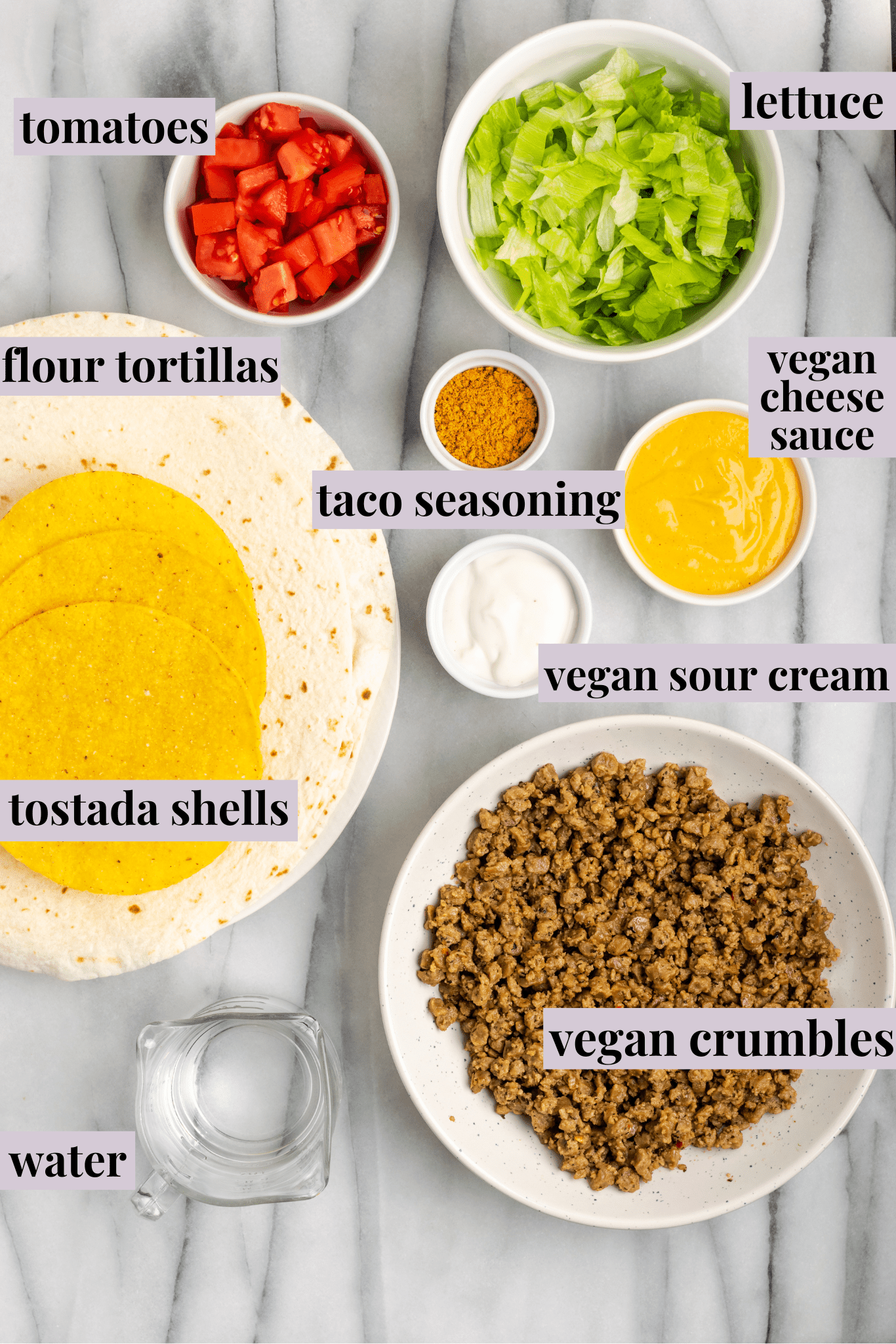 Overhead view of the ingredients for vegan crunchwrap supremes: flour tortillas, tostadas, a bowl of vegan meat crumbles, a bowl of lettuce, a bowl of tomatoes, a bowl of taco seasoning, a bowl of vegan sour cream, a bowl of vegan cheese sauce, and a glass of water