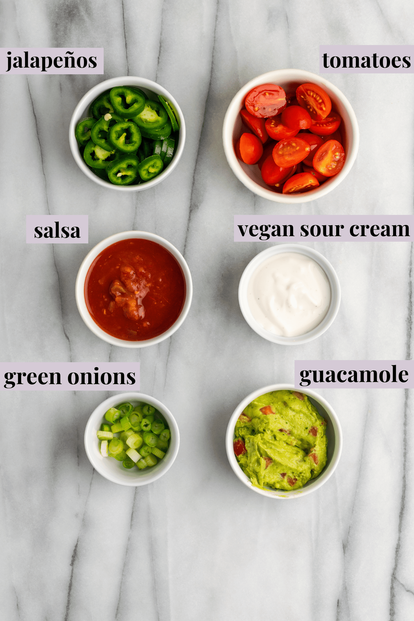 Overhead view of a bowl of jalapeño slices, a bowl of halved cherry tomatoes, a bowl of salsa, a bowl of vegan sour cream, a bowl of chopped scallions, and a bowl of guacamole