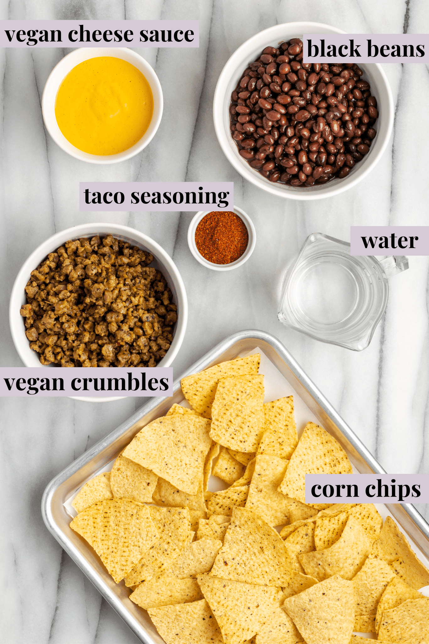 Overhead view of a tray of tortilla chips, a bowl of vegan meat crumble, a bowl of black beans, a bowl of vegan cheese sauce, a bowl of taco seasoning, and a glass of water
