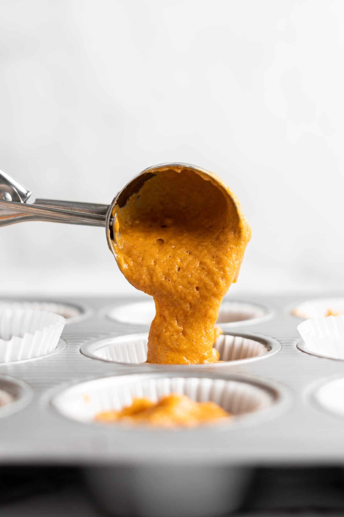 An ice cream scoop dropping pumpkin cupcake batter into a cupcake liner in a cupcake pan.