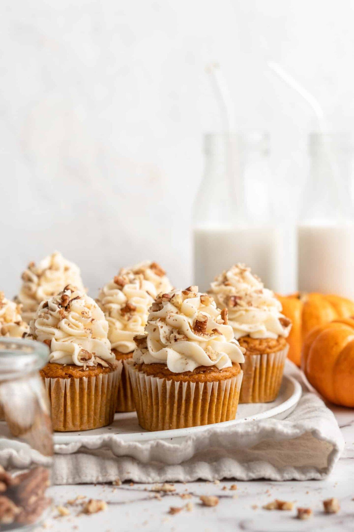 A plate full of pumpkin cupcakes topped with cream cheese frosting and pecans, with pumpkins and glasses of milk in the background.