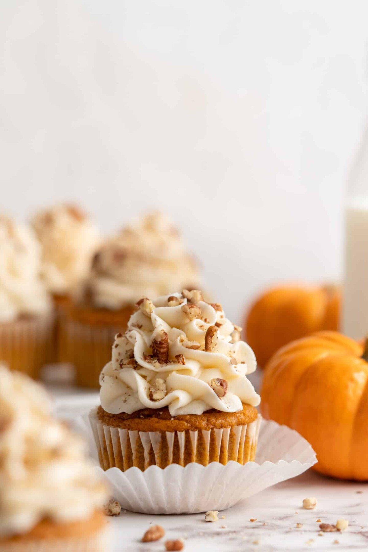 A pumpkin cupcake topped with cream cheese frosting and pecans, with more cupcakes and pumpkins in the background.