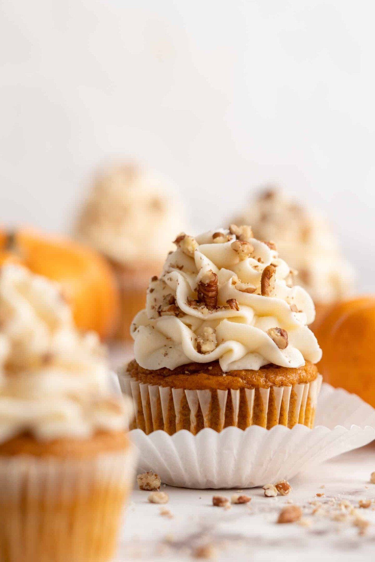 Close up of a pumpkin cupcake topped with cream cheese frosting and pecans, with more cupcakes and pumpkins in the background, and a cupcake in the foreground