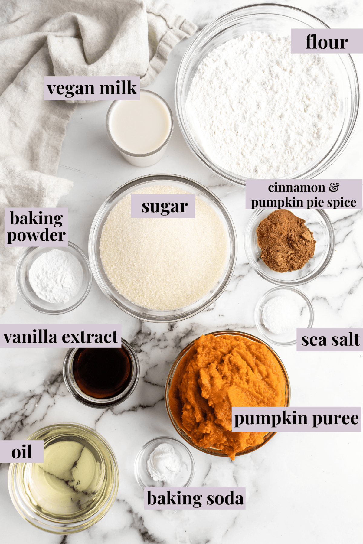 Overhead view of the labeled ingredients for pumpkin cupcakes: a bowl of flour, a bowl of sugar, a bowl of pumpkin puree, a bowl of vanilla extract, a bowl of sea salt, a bowl of baking powder, a bowl of baking soda, a bowl of vegan milk, a bowl of oil, and a bowl of cinnamon and pumpkin pie spice.