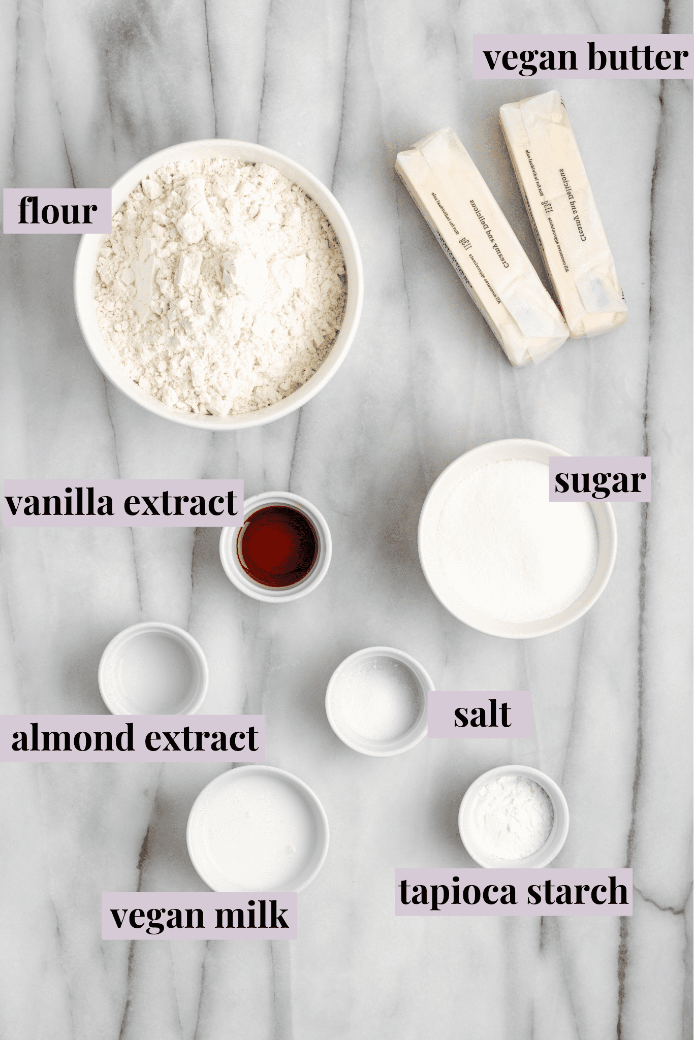 Overhead view of the ingredients for vegan spritz cookies: two sticks of vegan butter, a bowl of flour, a bowl of sugar, a bowl vegan milk, a bowl of cornstarch, a bowl of salt, a bowl of almond extract, and a bowl of vanilla extract