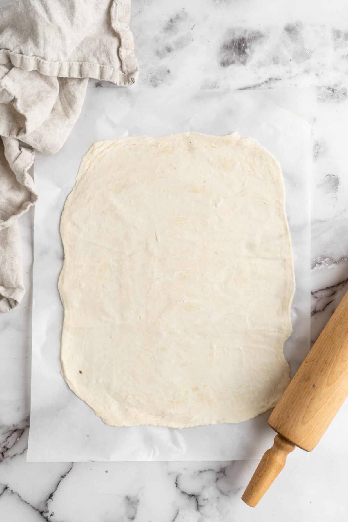 A sheet of puff pastry rolled into a rectangle, next to a kitchen towel and a rolling pin