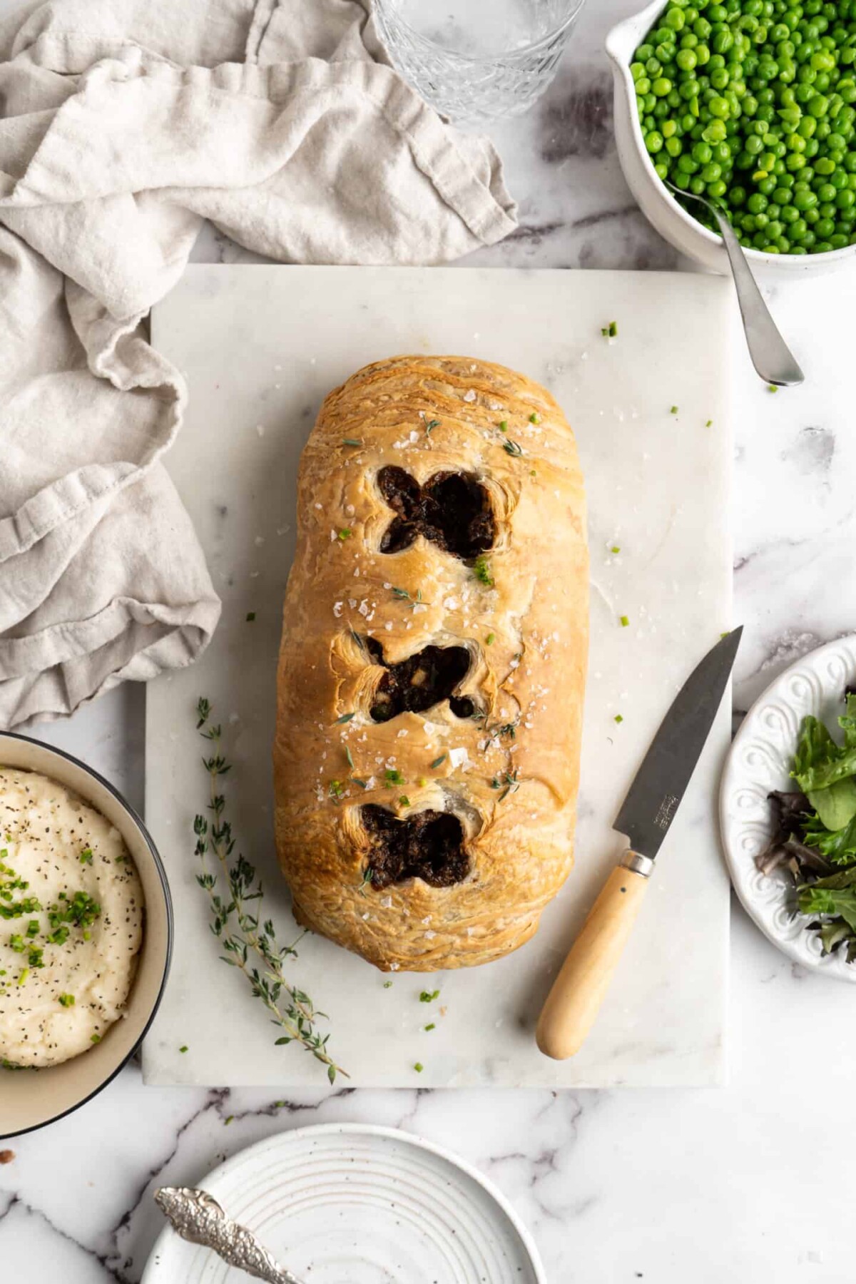 A vegan wellington with scores on the top, on a cutting board next to a paring knife and fresh thyme sprigs, a kitchen towel, a bowl of mashed potatoes, a bowl of peas, and a plate of greens