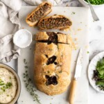 A vegan wellington loaf that's been cut into, with two slices on the cutting board, surrounded by a paring knife and some sprigs of time, next to a bowl of mashed potatoes