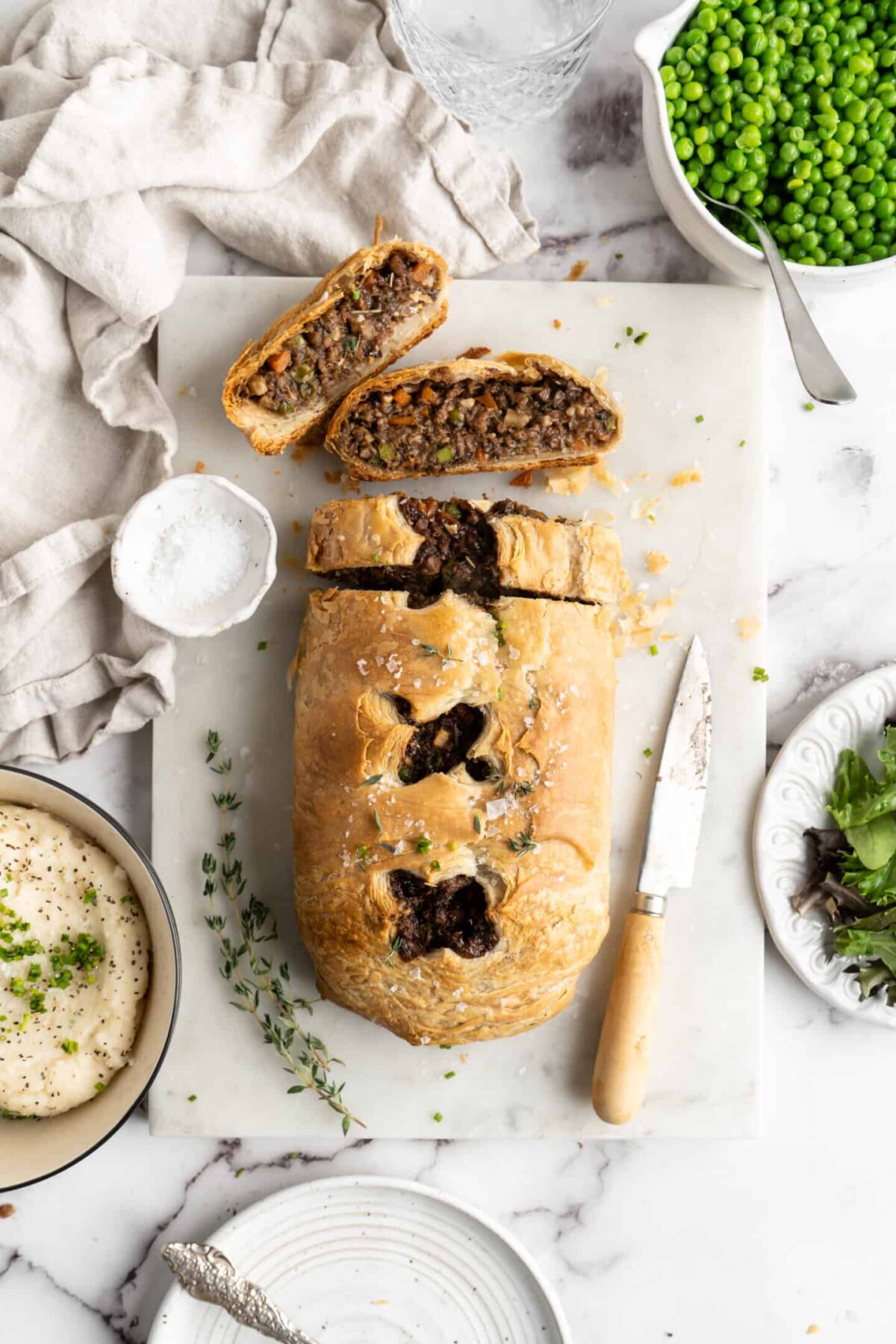 A vegan wellington loaf that's been cut into, with two slices on the cutting board, surrounded by a paring knife and some sprigs of time, next to a bowl of mashed potatoes