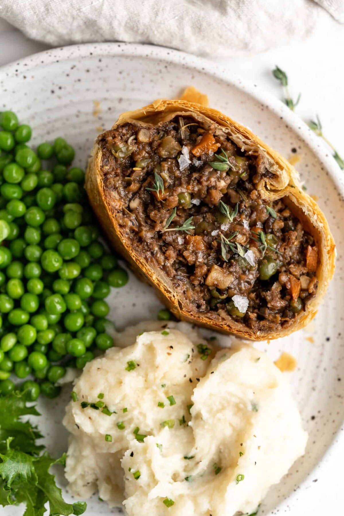 A slice of vegan wellington on a plate next to peas and mashed potatoes
