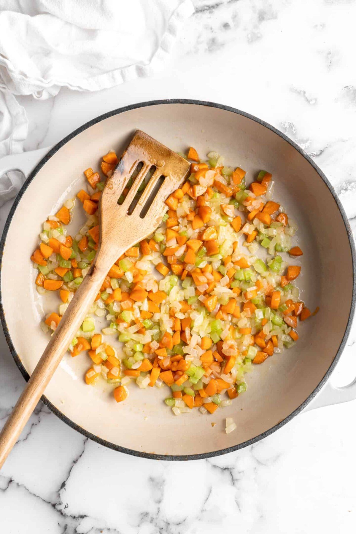 A pan with onions, celery, and carrots cooking in it, with a wooden spoon in the pan.