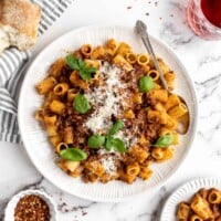 A plate of walnut lentil bolognese, topped with basil and vegan parmesan, with a fork on the plate, next to plates of pasta, a baguette, and a bowl of chili flakes