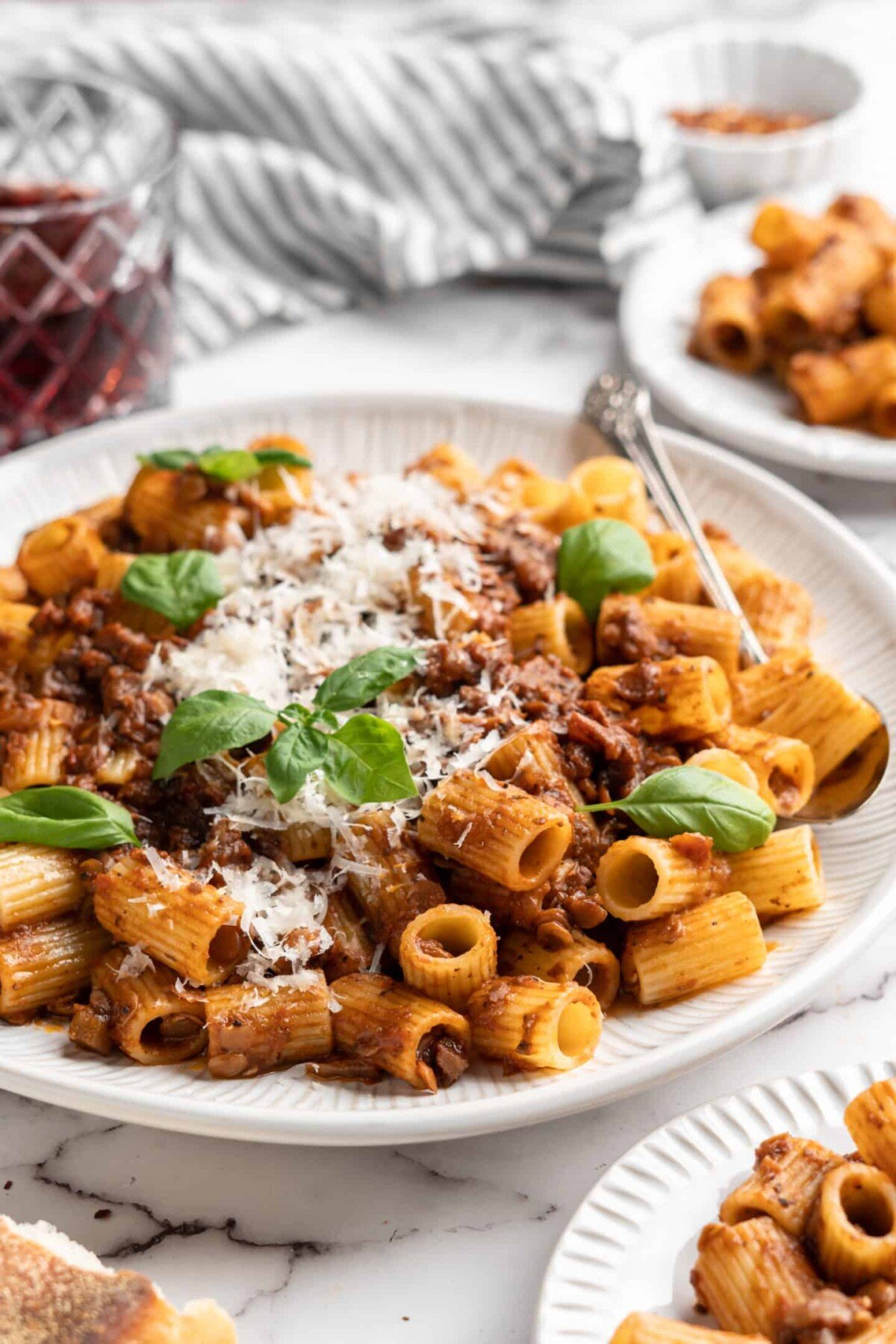 A plate of walnut lentil bolognese, topped with vegan parmesan and basil leaves, surrounded by more plates of pasta