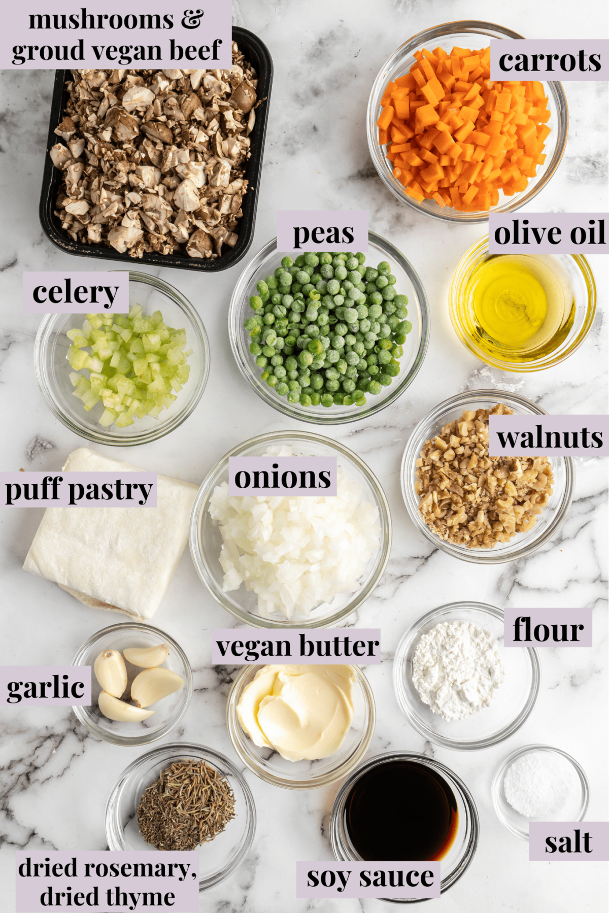 Overhead view of the ingredients for vegan wellingtons: a container of mushrooms and vegan beef, a bowl of carrots, a bowl of celery, a bowl of onions, a bowl of frozen peas, a bowl of walnuts, a bowl of flour, a bowl of olive oil, a bowl of garlic, a bowl of vegan butter, and a chunk of puff pastry