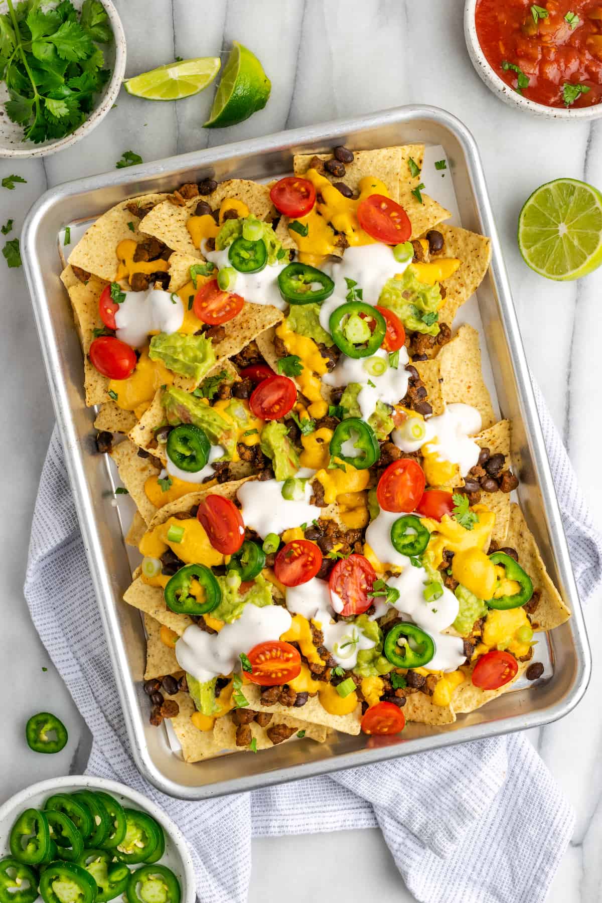 Overhead view of a tray of loaded nachos, covered in vegan sour cream, jalapeño slices, tomatoes, and cilantro, next to a half a lime, a bowl of cilantro, a bowl of jalapeños, and a bowl of salsa