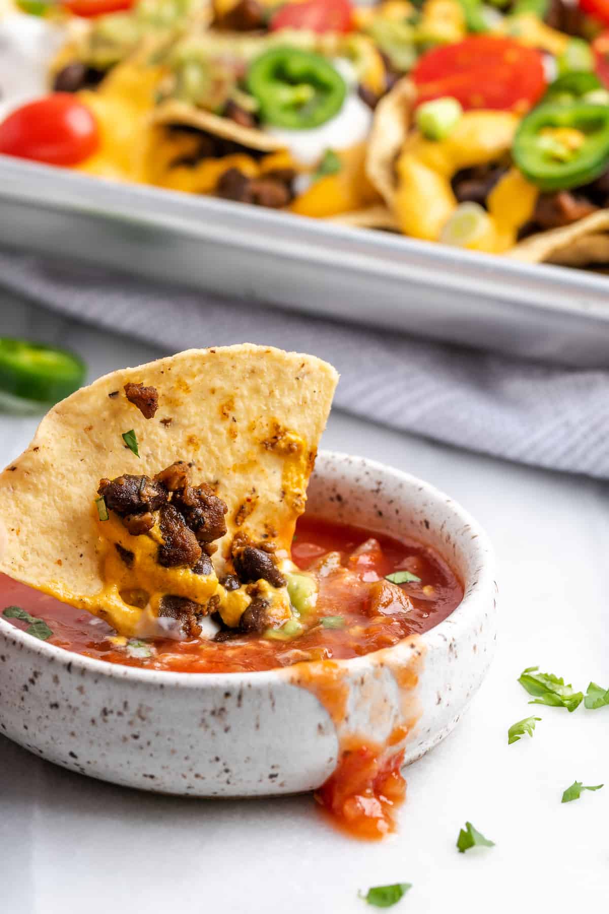 A chip with beans and cheese on it dipping into a bowl of salsa, with a tray of nachos in the background