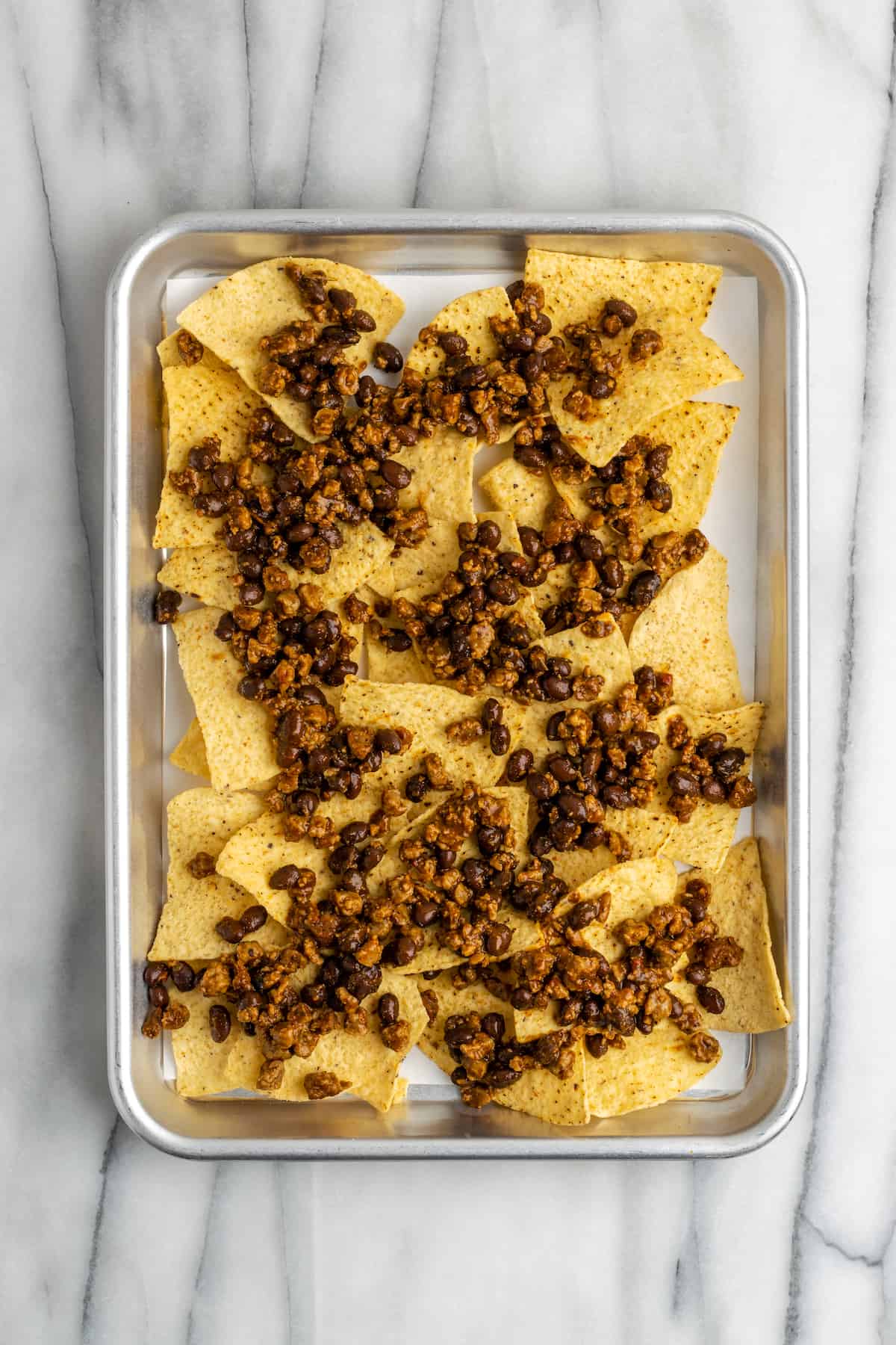 A baking tray with a layer of tortilla chips, topped with a mixture of black beans and vegan meat crumbles