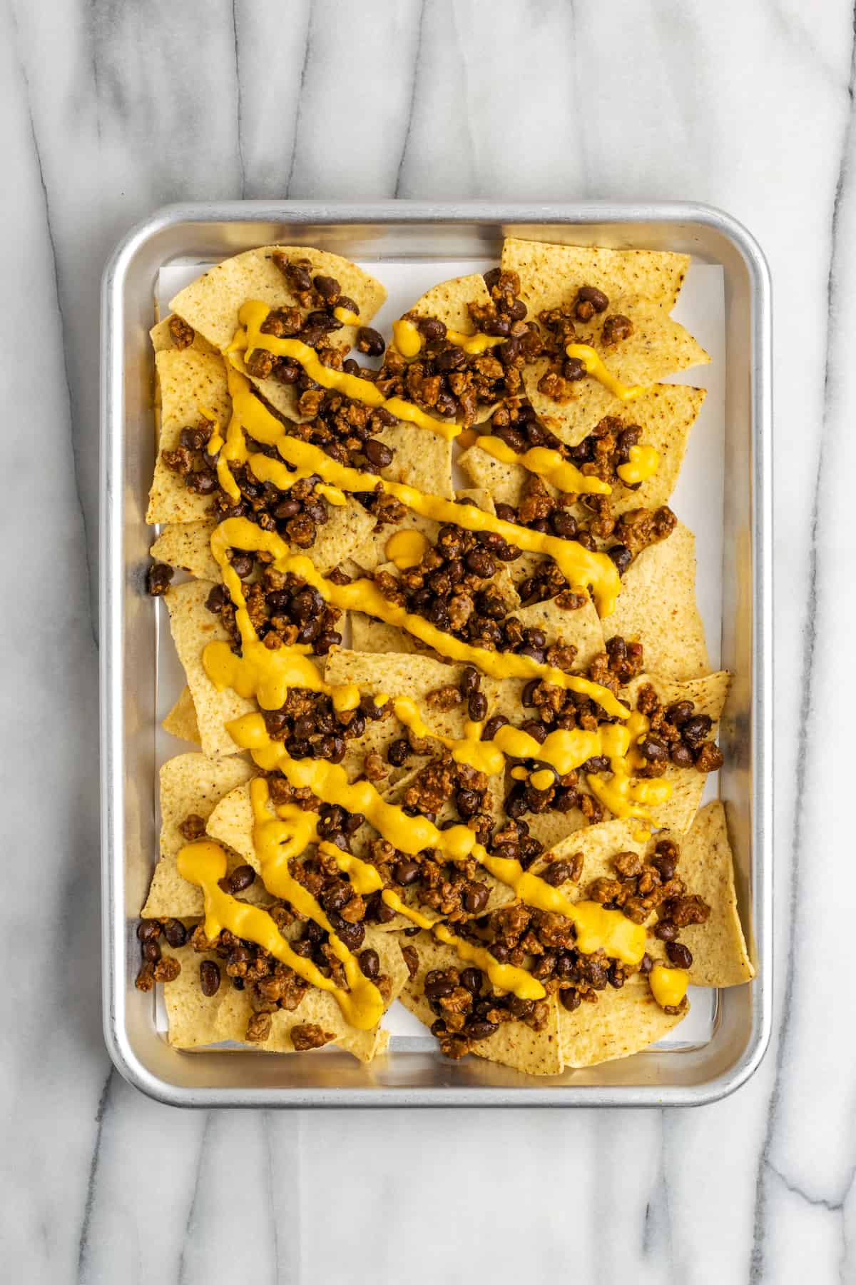 A baking sheet with a layer of chips, topped with vegan meat crumbles and beans, with vegan cheese sauce poured on top