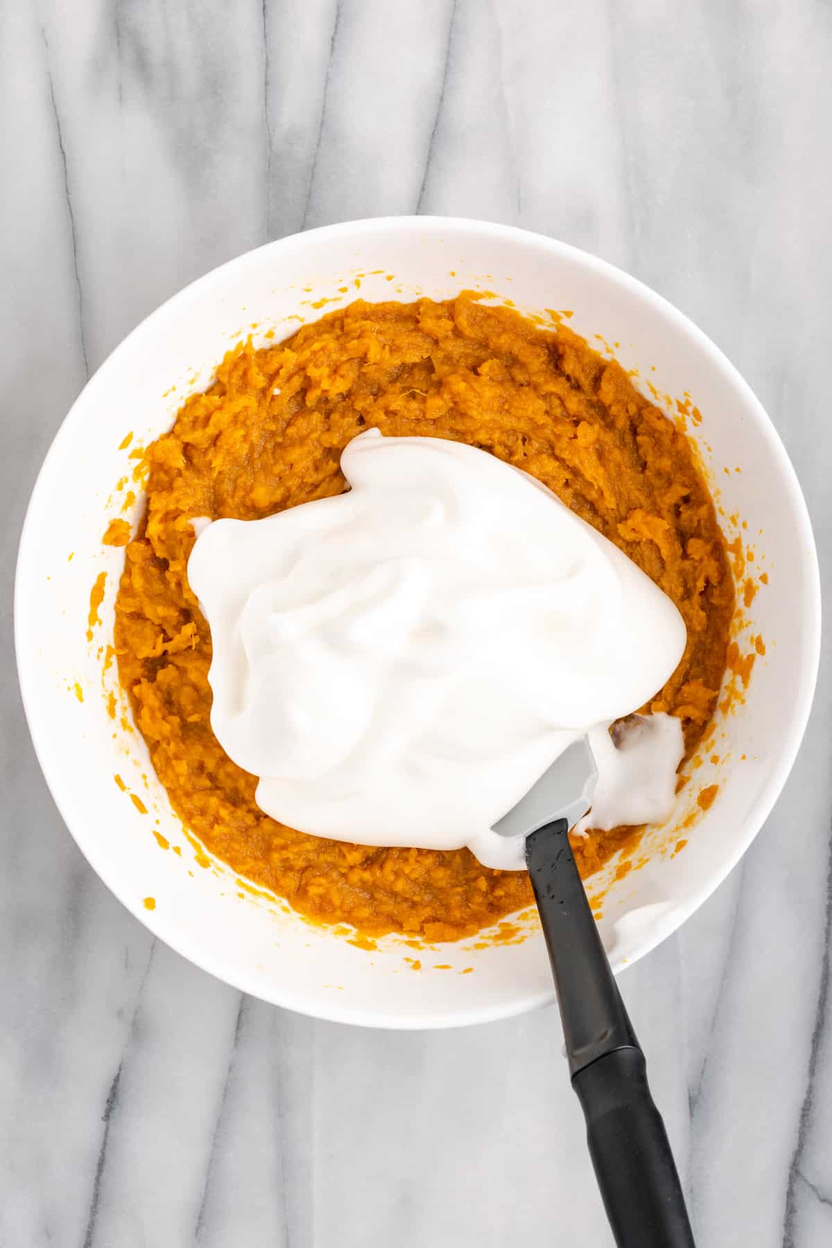 A mixing bowl with whipped aquafaba sitting on top of sweet potato soufflé batter, with a spatula in the bowl