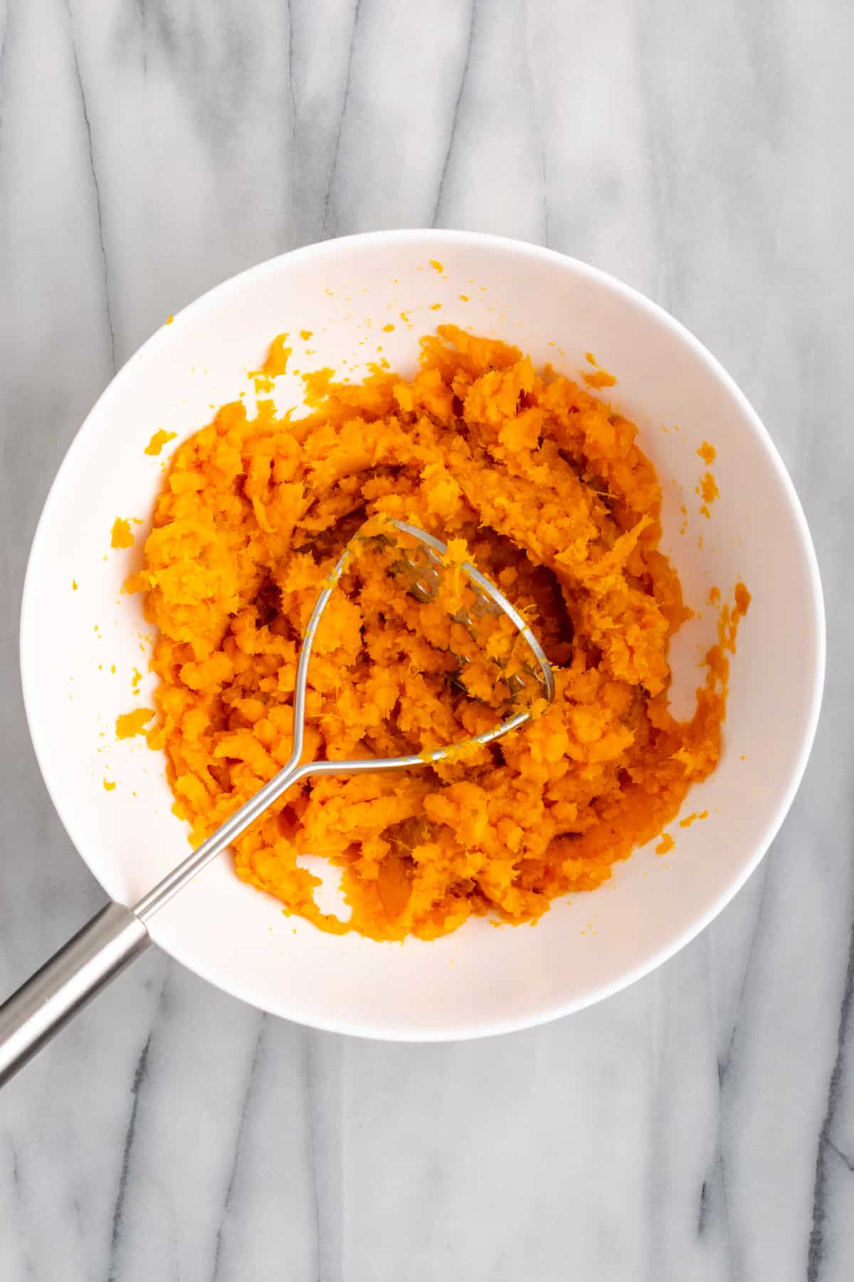 Mashed sweet potatoes in a mixing bowl with a potato masher in it