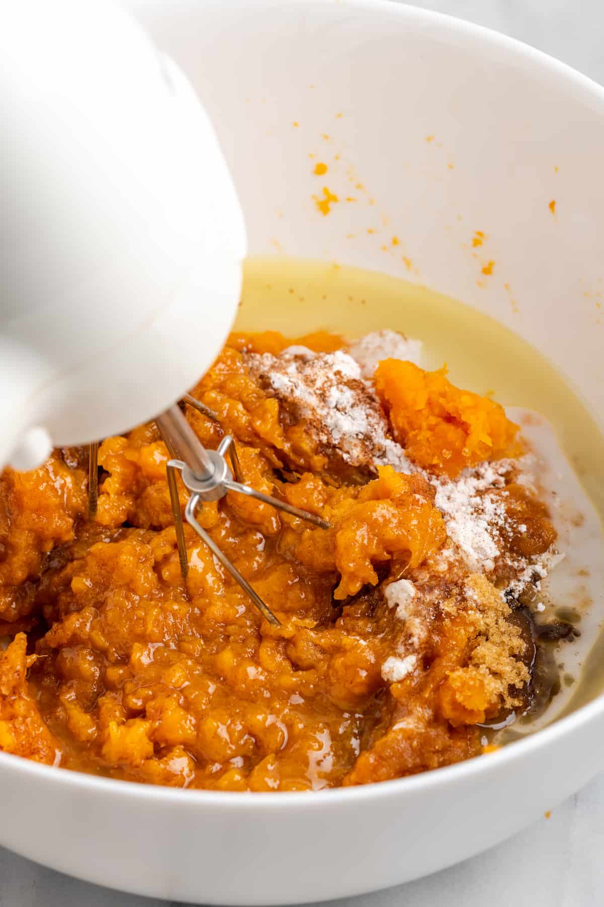 A hand mixer whipping together sweet potatoes with maple syrup, vegan butter, non-dairy milk, and seasonings, in a mixing bowl.