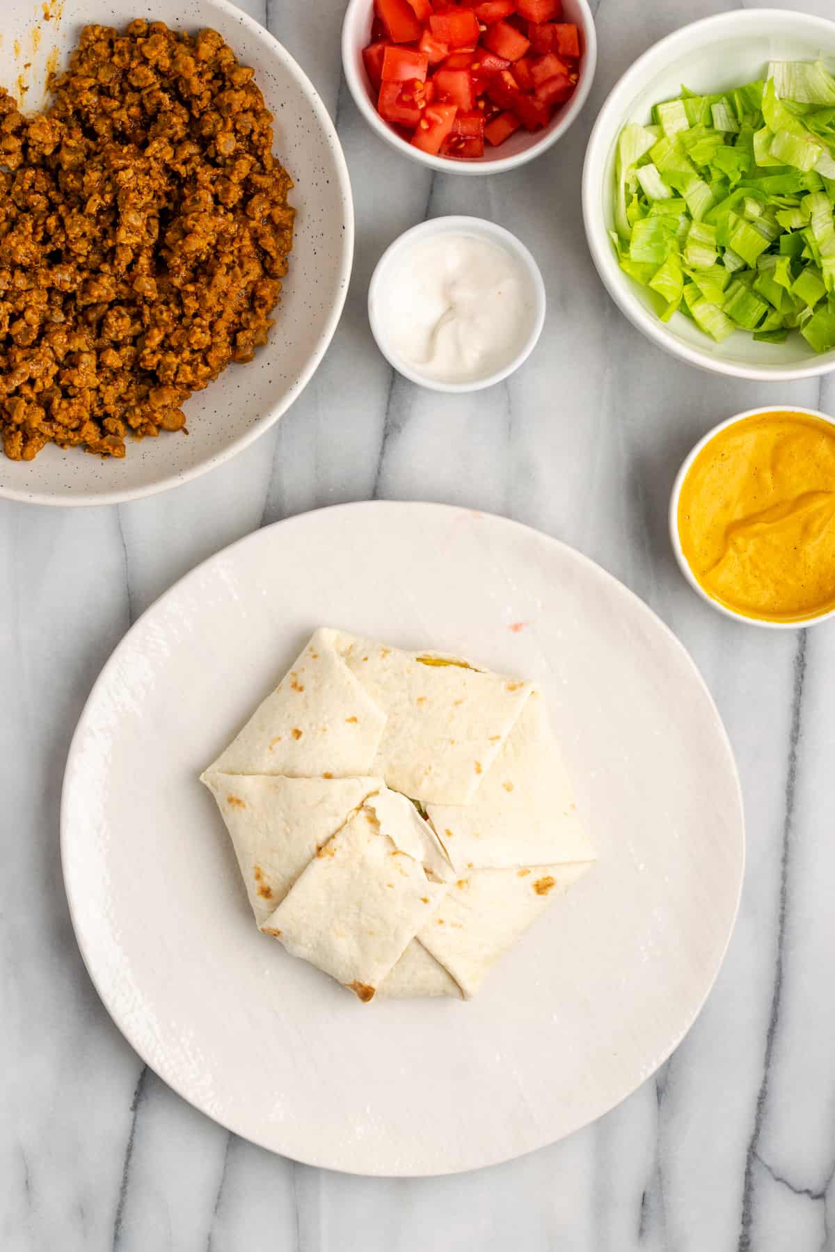 A plate with an untoasted crunchwrap on it, folded up, next to a bowl of vegan meat crumbles, a bowl of vegan sour cream, a bowl of vegan cheese sauce, a bowl of lettuce, and a bowl of tomatoes.