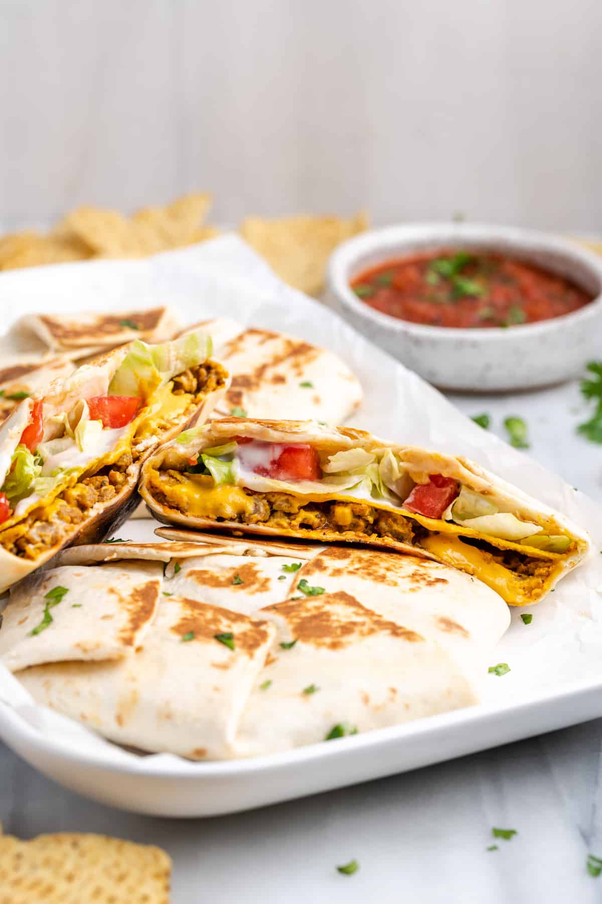 A rectangular plate with a vegan crunchwrap supreme cut in half on top of other crunchwraps, with a bowl of salsa in the background