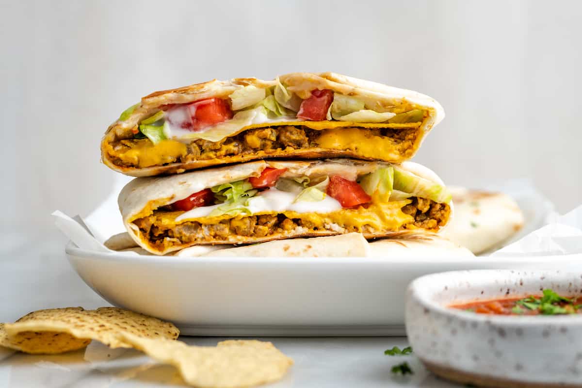 A vegan crunchwrap supreme cut in half, with the two sides on top of each other, cut side facing forward, on top of a whole crunchwrap supreme, surrounded by tortilla chips